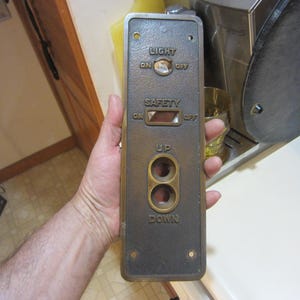 May include: A vintage elevator control panel with brass accents. The panel has buttons and switches labeled "LIGHT", "SAFETY", "UP", and "DOWN". The panel is rectangular and has a dark, aged finish.