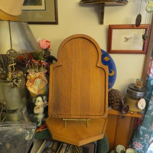 May include: A wooden cutting board with a decorative arched top and a stand. The board is made of light brown wood and has a smooth surface. The stand is made of metal and has a gold finish.