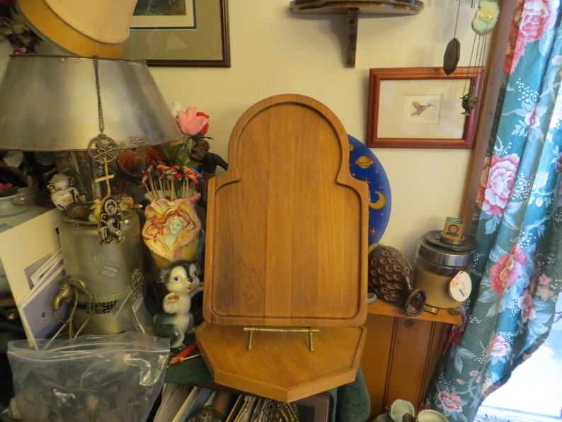 May include: A wooden cutting board with a decorative arched top and a stand. The board is made of light brown wood and has a smooth surface. The stand is made of metal and has a gold finish.
