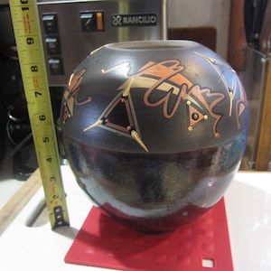 May include: A black ceramic vase with a geometric design in gold, copper, and white. The vase has a round shape and a glossy finish.