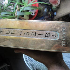May include: A vintage brass doorbell with the word "TRIBORO" engraved on the side. The doorbell features a series of perforated dots and a black button. The brass has a worn, aged appearance.