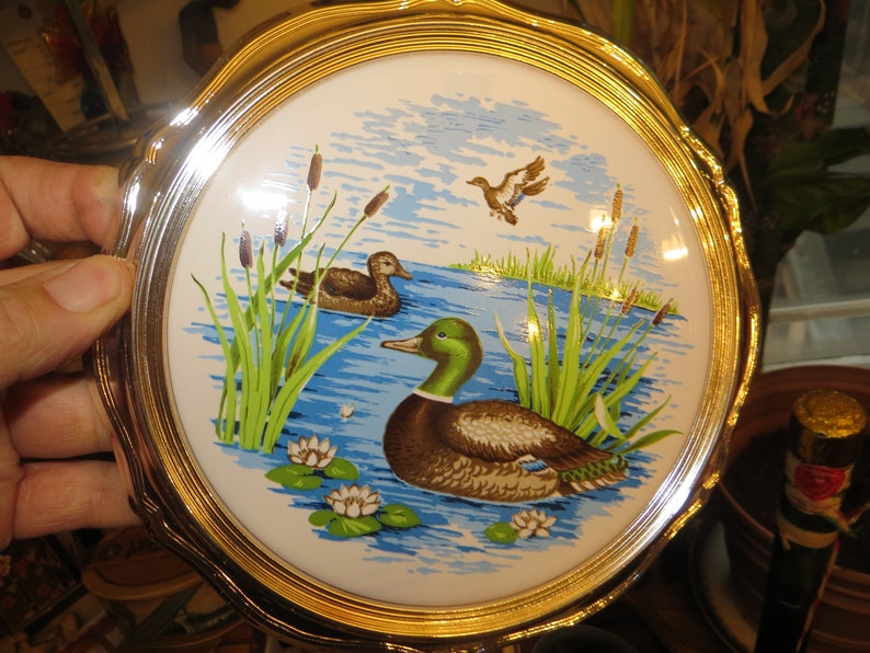 Puede incluir: Un plato redondo de cer&aacute;mica blanca con un borde dorado presenta una escena pintada de dos patos nadando en un estanque con juncos verdes y nen&uacute;fares blancos. El plato es perfecto para exhibir o servir aperitivos.