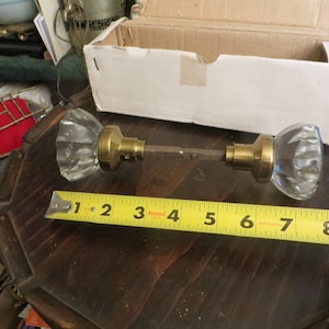 May include: A pair of vintage crystal door knobs with brass hardware. The knobs are approximately 6 inches long, with a clear, faceted design. A yellow measuring tape is placed next to the knobs.