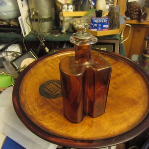 May include: A brown glass bottle with a unique shape and a silver metal stopper. The bottle is sitting on a wooden tray with a dark brown rim.