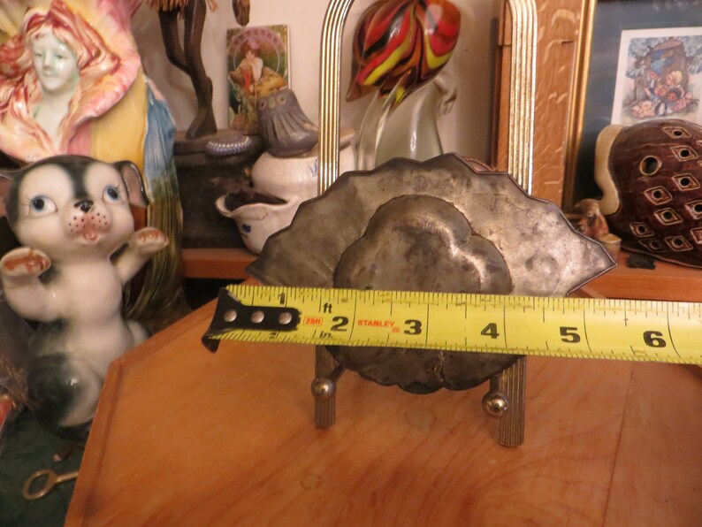 May include: A silver-toned metal napkin holder with a decorative design. The holder is 10 cm wide and is sitting on a wooden surface.