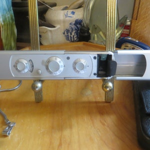 May include: A silver spy camera with three dials, open and showing the inside of the camera. The camera is on a wooden surface with a silver chain and a black leather case.