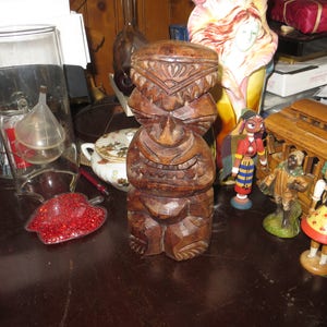 May include: A wooden tiki statue with a carved face and a detailed design. The statue is brown and has a dark wood finish.