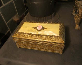 May include: A gold-colored jewelry box with an ornate design. The top of the box is made of a light yellow material with a pink cameo in the center.