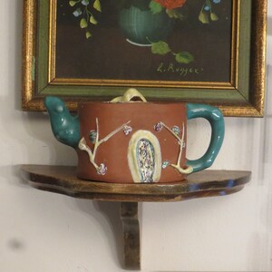 May include: A brown teapot with a green spout and handle. The teapot is decorated with a painted design of branches and flowers. The teapot is sitting on a wooden shelf.
