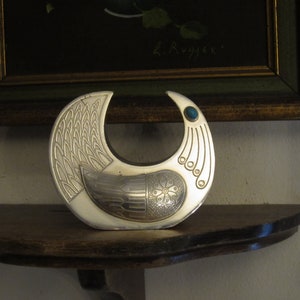 May include: A silver-toned, crescent-shaped decorative object with intricate embossed patterns. The design includes a bird-like motif with a teal-colored stone accent. It rests on a dark wooden shelf, with a framed painting in the background.