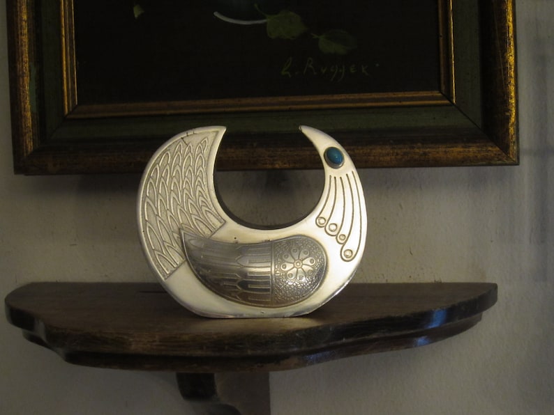 May include: A silver-toned, crescent-shaped decorative object with intricate embossed patterns. The design includes a bird-like motif with a teal-colored stone accent. It rests on a dark wooden shelf, with a framed painting in the background.