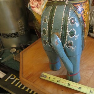 Puede incluir: Una figurita de elefante de madera azul con detalles pintados, incluyendo puntos blancos, c&iacute;rculos verdes y rojos, y patrones florales amarillos y naranjas. El elefante est&aacute; de pie sobre una base de madera con una cinta m&eacute;trica.