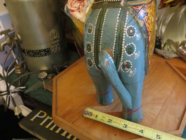 Puede incluir: Una figurita de elefante de madera azul con detalles pintados, incluyendo puntos blancos, c&iacute;rculos verdes y rojos, y patrones florales amarillos y naranjas. El elefante est&aacute; de pie sobre una base de madera con una cinta m&eacute;trica.