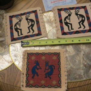 May include: Three square woven coasters with a Native American design featuring two figures playing flutes. The coasters are beige with a red, blue, and black design. The figures are black with white accents. The coasters are approximately 4 inches by 4 inches.