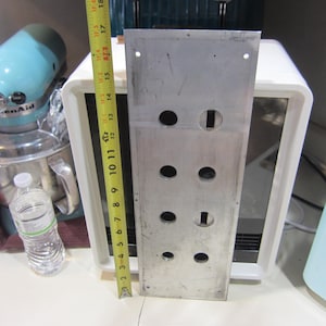 May include: A rectangular, silver-colored metal plate with several circular and rectangular holes. A yellow measuring tape shows the plate is approximately 18 inches long. A KitchenAid mixer and other kitchen items are in the background.