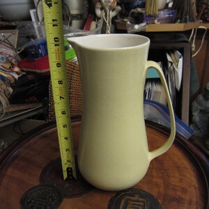 May include: A pale yellow ceramic pitcher with a white interior, approximately 11 inches tall. The pitcher has a curved handle and a spout for pouring. A yellow measuring tape is placed next to the pitcher.