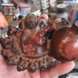 May include: A brown ceramic figurine of a person with a smiling face and a red and white patterned dress. The figurine is holding a brown ceramic pot with red and white patterns.