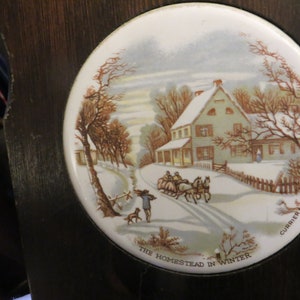 May include: A white ceramic tile with a winter scene featuring a house with a snow-covered roof and a horse-drawn carriage. The tile is decorated with a brown wooden frame and the text "The Homestead in Winter" and "Currier & Ives".