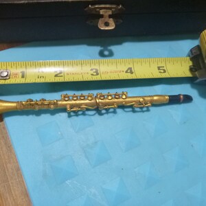 May include: A gold-colored miniature clarinet measuring approximately 5 inches long. The clarinet has a black mouthpiece and is resting on a blue surface.