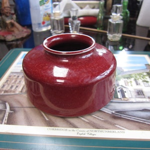 May include: A deep red ceramic vase with a white rim. The vase has a glossy finish and is sitting on a green and white placemat with a picture of a village.