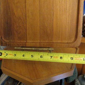 May include: A wooden cutting board with a brass hinge and a yellow measuring tape showing 10 inches. The cutting board is made of light brown wood and has a rectangular shape.