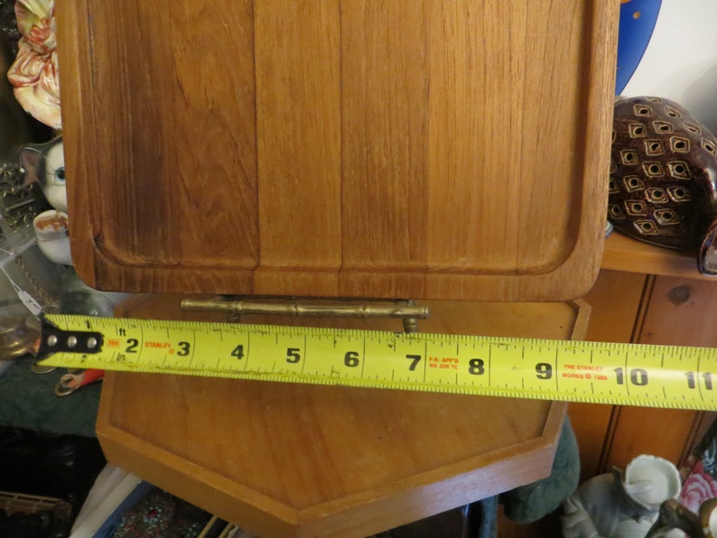 May include: A wooden cutting board with a brass hinge and a yellow measuring tape showing 10 inches. The cutting board is made of light brown wood and has a rectangular shape.