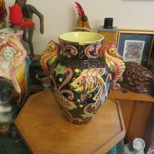 May include: A black ceramic vase with a yellow rim and gold accents. The vase is decorated with a seahorse design on each side and a fish design in the center. The vase is painted with pink, yellow, and green colors.