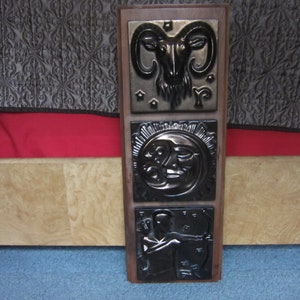 May include: A wooden wall hanging with three black and silver ceramic tiles. The top tile features a ram's head, the middle tile features a sun and moon, and the bottom tile features a figure with a bow and arrow.