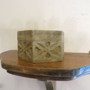 May include: A small, hexagonal, carved stone box with intricate floral designs. The box is light brown in color and has a smooth, polished finish. It is sitting on a wooden shelf.