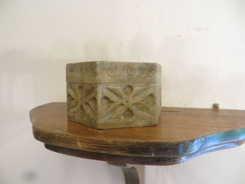 May include: A small, hexagonal, carved stone box with intricate floral designs. The box is light brown in color and has a smooth, polished finish. It is sitting on a wooden shelf.