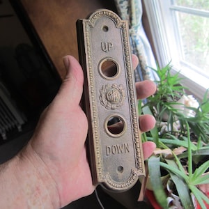 May include: Vintage brass elevator call button plate with the words "UP" and "DOWN" and two circular button holes. The plate has an ornate design with a decorative emblem in the center. The plate is held in a hand.