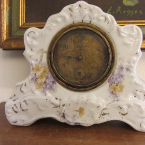 May include: White porcelain clock with a gold-trimmed face and floral accents. The clock has a Roman numeral dial and is set on a wooden surface.