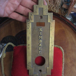 May include: A brass door plate with a rectangular shape and an Art Deco design. The plate has a raised inscription that reads "KIMBALL".