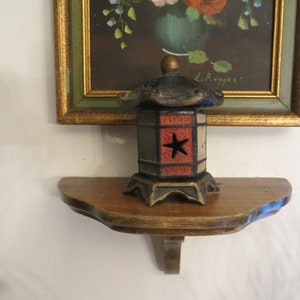 May include: A small, black, red, and green cast iron lantern with a star design sits on a wooden shelf. The lantern has a pagoda-style roof and is decorated with a red star on the front. The shelf is attached to a white wall.