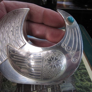 May include: A silver-toned, crescent-shaped brooch with intricate engraved feather and floral patterns. A small, round turquoise stone accents one end. The brooch has a detailed, artistic design.