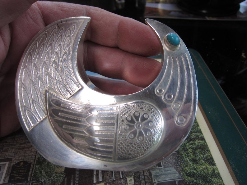 May include: A silver-toned, crescent-shaped brooch with intricate engraved feather and floral patterns. A small, round turquoise stone accents one end. The brooch has a detailed, artistic design.