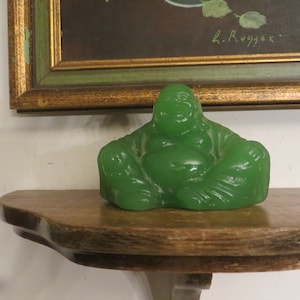 May include: A green jade figurine of a smiling Buddha sitting in a meditative pose. The figurine is on a wooden shelf.