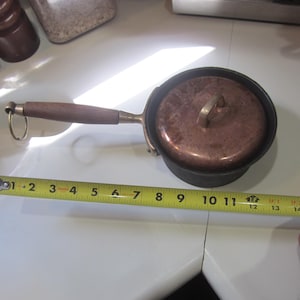 May include: A small, vintage copper saucepan with a lid, wooden handle, and brass accents. The pan measures approximately 11 inches in length, including the handle. The lid has a brass loop handle.