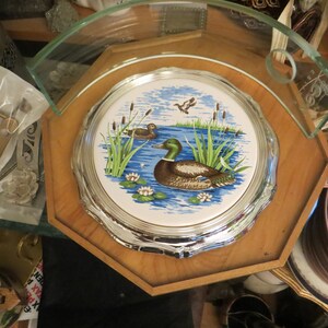 Puede incluir: Una bandeja octogonal de madera con un posavasos de cer&aacute;mica con borde plateado que representa una escena de lago azul con dos patos y juncos verdes. El posavasos est&aacute; rodeado por un borde plateado.