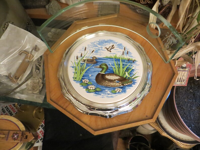 Puede incluir: Una bandeja octogonal de madera con un posavasos de cer&aacute;mica con borde plateado que representa una escena de lago azul con dos patos y juncos verdes. El posavasos est&aacute; rodeado por un borde plateado.