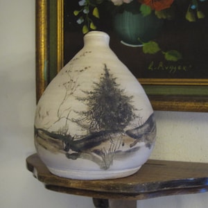 May include: A white ceramic vase with a hand-painted design of a forest scene. The vase has a rounded body and a narrow neck. The design features a tall pine tree, smaller trees, and a path through the woods. The vase is sitting on a wooden shelf.