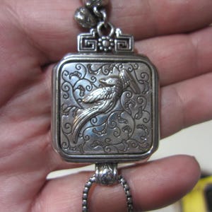 Pu&ograve; includere: Ciondolo argentato con un design rettangolare inciso raffigurante un uccello e motivi floreali. Il ciondolo &egrave; sospeso da una catena e ha un elemento superiore decorativo. Il ciondolo &egrave; tenuto in una mano.