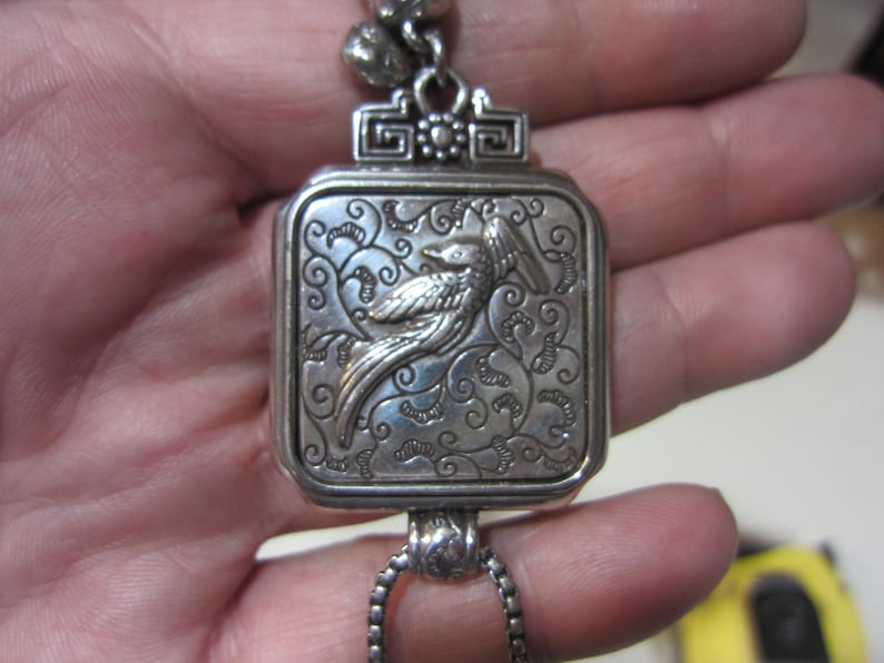 Pu&ograve; includere: Ciondolo argentato con un design rettangolare inciso raffigurante un uccello e motivi floreali. Il ciondolo &egrave; sospeso da una catena e ha un elemento superiore decorativo. Il ciondolo &egrave; tenuto in una mano.