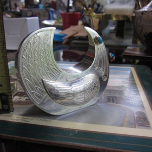 May include: A silver-colored bird-shaped bracelet with etched feather and wing details. The bracelet has a turquoise stone accent and measures approximately 2 inches in diameter.