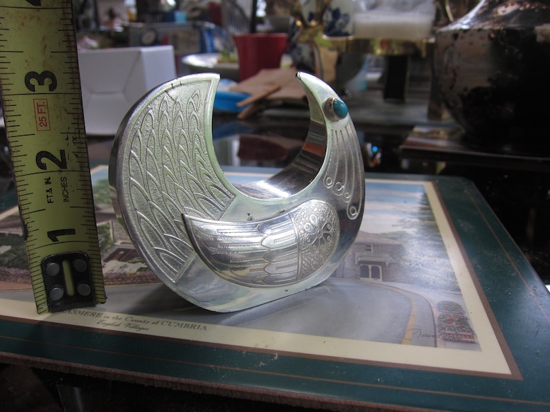 May include: A silver-colored bird-shaped bracelet with etched feather and wing details. The bracelet has a turquoise stone accent and measures approximately 2 inches in diameter.