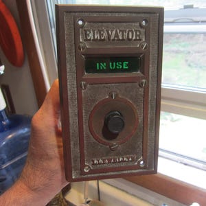 May include: A rectangular, bronze-colored elevator control panel with the word "ELEVATOR" at the top. A green display reads "IN USE". The panel has a circular dial and is secured with screws.