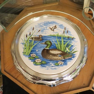 Puede incluir: Un posavasos de cer&aacute;mica con borde plateado, decorado con un dise&ntilde;o azul y blanco que representa dos patos nadando en un estanque con juncos verdes y nen&uacute;fares blancos.