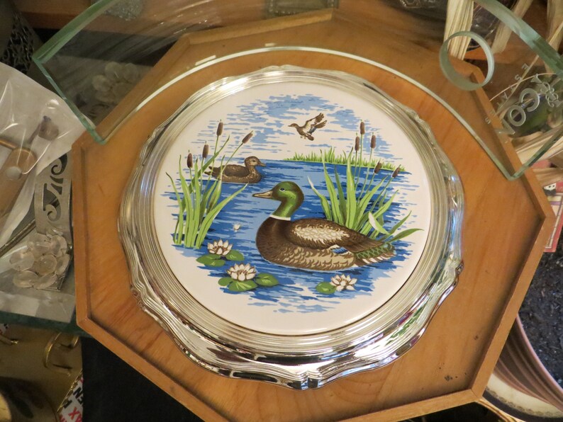 Puede incluir: Un posavasos de cer&aacute;mica con borde plateado, decorado con un dise&ntilde;o azul y blanco que representa dos patos nadando en un estanque con juncos verdes y nen&uacute;fares blancos.