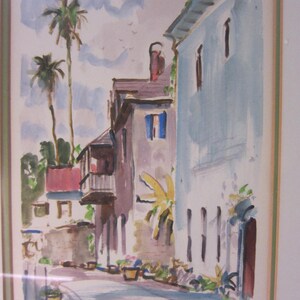 May include: Watercolor painting of a street scene with buildings, palm trees, and a blue sky. The buildings are painted in shades of pink, blue, and white. The palm trees are green and the sky is a light blue. The painting is signed "Ernest Duffy" in the bottom right corner.