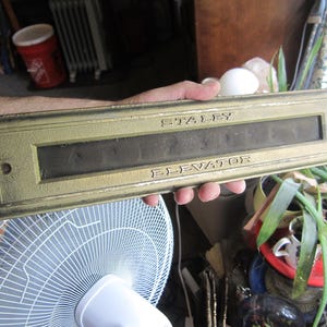 May include: Vintage brass elevator indicator plate with the text "STALEY ELEVATOR". The rectangular plate has a dark, recessed window and is held in a person's hand. The plate has a gold finish with some wear.